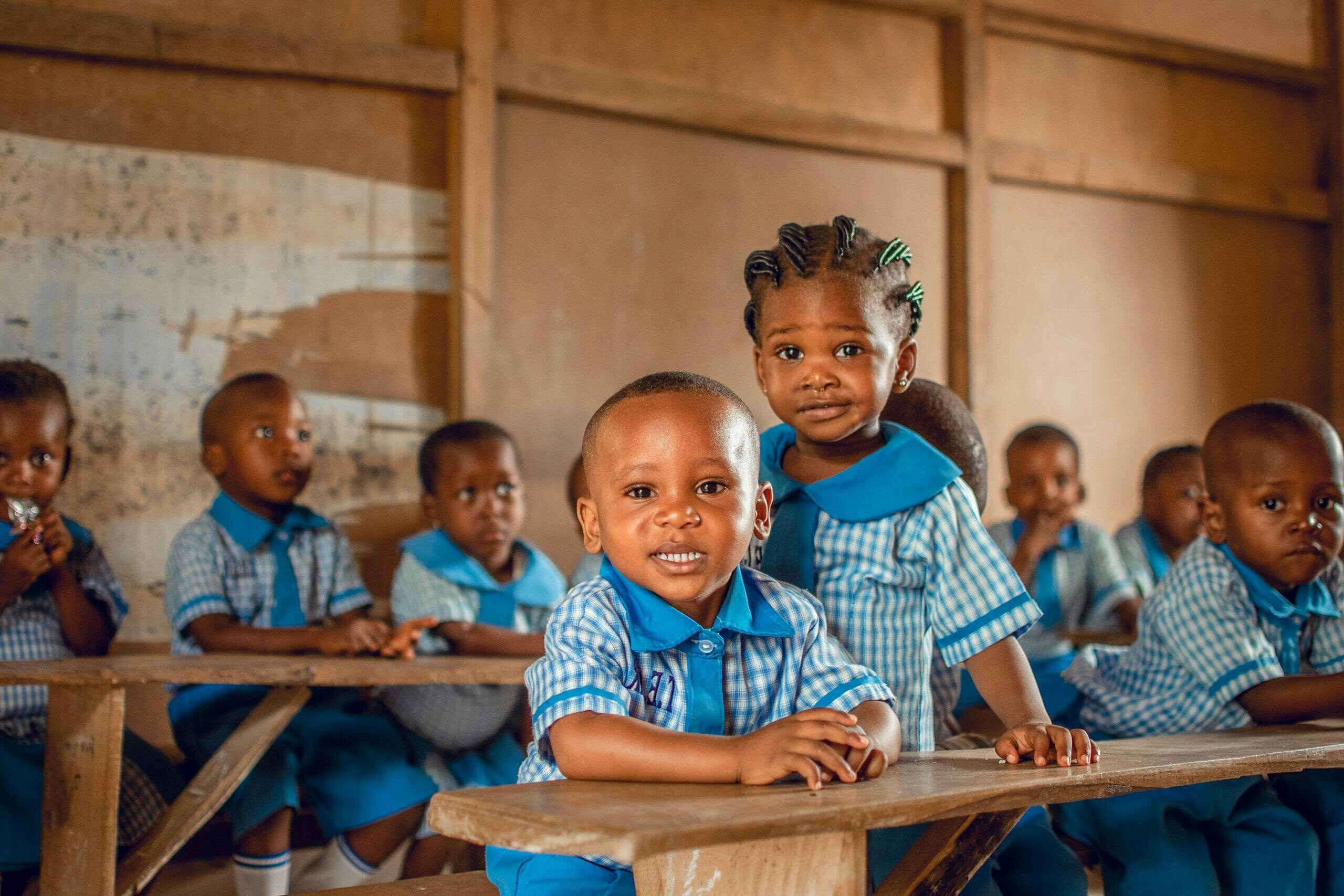 Happy children in a Nigerian classroom wearing uniforms, sitting on wooden benches.