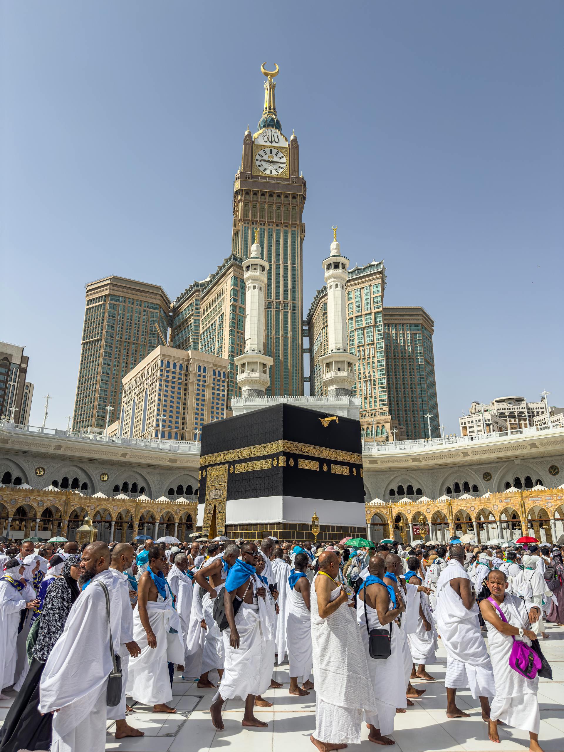 A large group of pilgrims gather at the Kaaba in Mecca, Saudi Arabia, during the Hajj pilgrimage under a clear blue sky.
