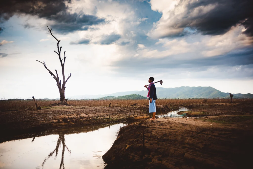 A farmer sits alone on cracked, dry earth beside a drying water source